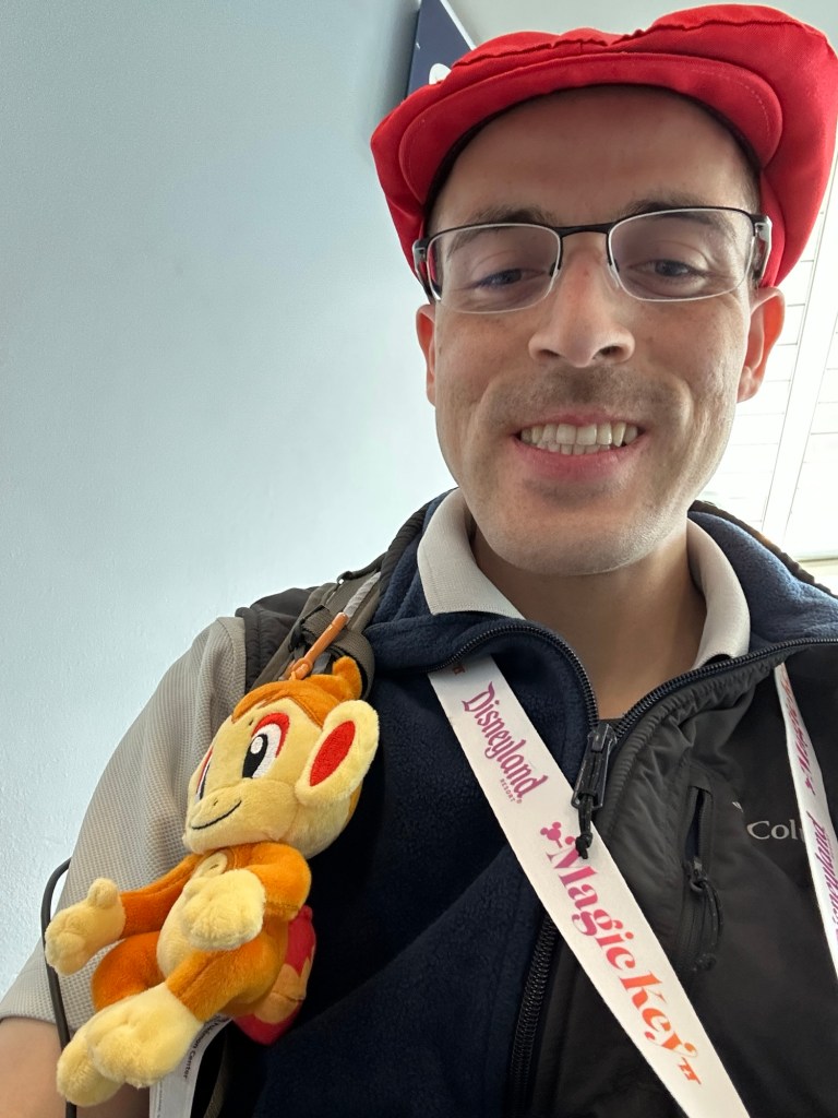 Anthony Solhtalab, cosplaying as Lucas from Pokémon Diamond & Pearl with a red flat hat, light gray polo beneath a navy polar fleece vest. He has a yellow backpack with a clip on plush of Chimchar, the fire Pokémon from Diamond & Pearl.