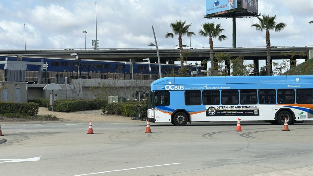 At the Anaheim Regional Transportation Intermodal Center (ARTIC) an Amtrak Pacific Surfliner departs in the background, and an OCBus arrives to the bus bays the midground.